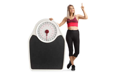 Full length portrait of a young woman in sportswear leaning on a big weight scale and pointing up isolated on white backgroundの写真素材