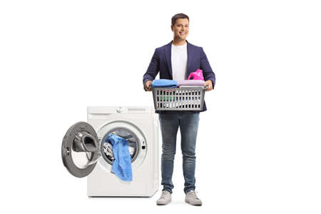 Young man carrying a laundry basket with clothes in front of a washing machine isolated on white backgroundの写真素材