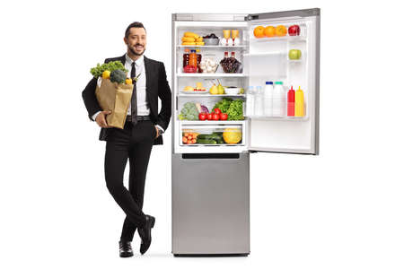 Full length portrait of a young businessman with a grocery bag leaning on a fridge isolated on white backgroundの写真素材