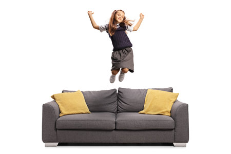 Happy schoolgirl in a uniform jumping on a sofa isolated on white backgroundの写真素材