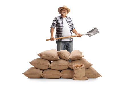 Mature farmer holding a shovel and posing with a pile of burlap sacks isolated on white background