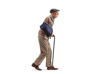 Full length profile shot of an elderly man with an arm splint walking with a cane isolated on white...