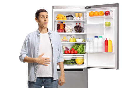 Young man holding his hand on his stomach in front of an open fridge with food isolated on white backgroundの写真素材