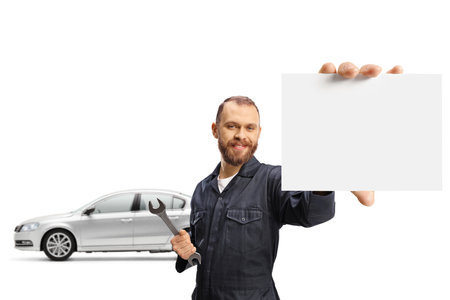 Mechanic posing in front of a car and holding a wrench and a small blank card isolated on white backgroundの写真素材
