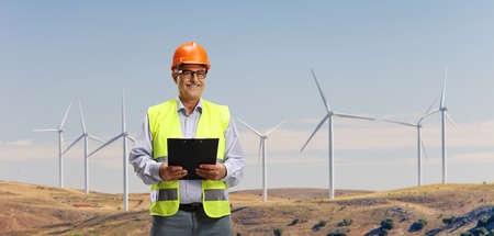Smiling mature engineer standing on a wind turbine farm and holding a clipboard