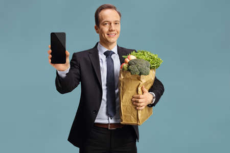 Bussinesman holding a paper bag with groceries and showing a smartphone isolated on blue backgroundの写真素材