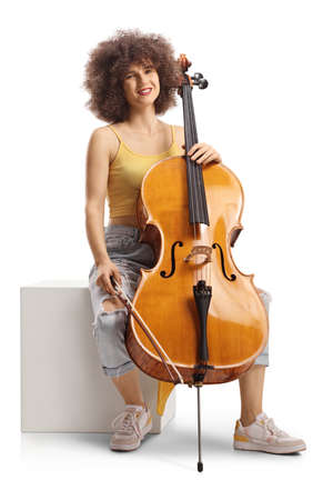 Young female musician sitting on a white cube with a cello and smiling isolated on white backgroundの写真素材