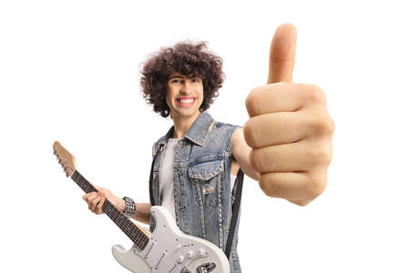 Smiling young male in a denim vest with an electric guitar showing thumbs up isolated on white...