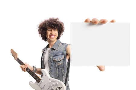 Young male in a denim vest standing with an electric guitar and showing a blank card isolated on...