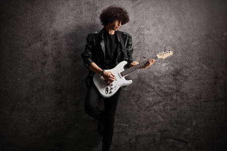 Male musician in a leather jacket with an electric guitar leaning on a dark rugged wallの写真素材