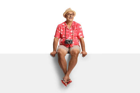 Happy mature male tourist with camera hanging around his neck sitting on a blank panel isolated on...