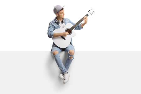 Guy sitting on a blank panel and playing an acoustic guitar isolated on white backgroundの写真素材