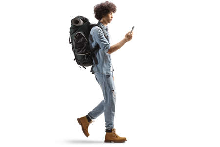 Full length profile shot of a young male backpacker walking and using a smartphone isolated on white...
