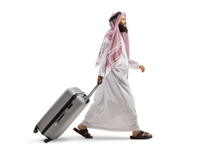 Full length profile shot of a saudi arab man walking and pulling a suitcase isolated on white backgroundの写真素材