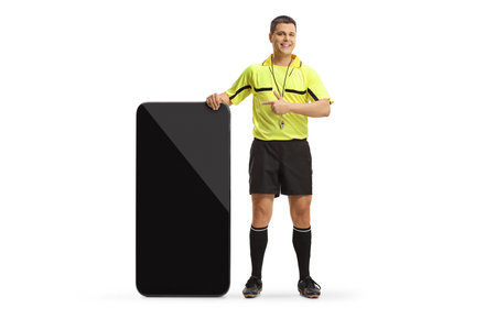 Full length portrait of a football referee standing next to a big mobile phone and pointing isolated on white backgroundの写真素材