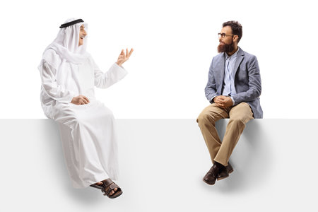 Arab man in an ethinc robe talking to a casual bearded man seated on a white board isolated on white backgroundの写真素材