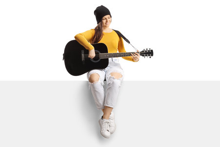 Full length portrait of a young trendy female playing an acoustic guitar and sitting on a blank panel isolated on white backgroundの写真素材