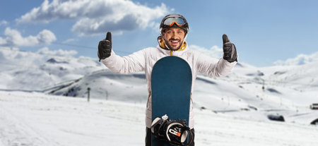 Young man with a snowboard standing on a mountain and gesturing with both thumbs upの写真素材