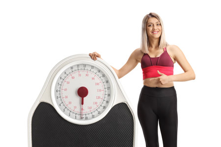 Young sporty woman leaning on a big weight scale and pointing isolated on white backgroundの写真素材