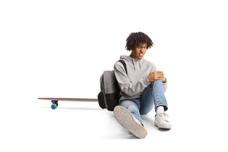 African american guy with a skateboard sitting on the ground and holding his injured knee isolated on white backgroundの写真素材