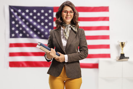 Young female teacher with glasses holding books with USA flag in the backgroundの写真素材