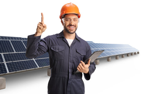 Young male factory worker standing in front of solar panels and pointing up isolated on white...