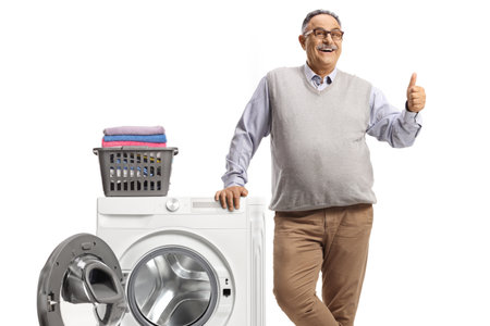 Mature man leaning on a washing machine with folded clothes and showing thumbs up isolated on white backgroundの写真素材