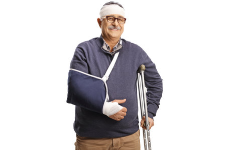 Mature man with a broken arm and bandage on head leaning on a crutch isolated on white backgroundの写真素材