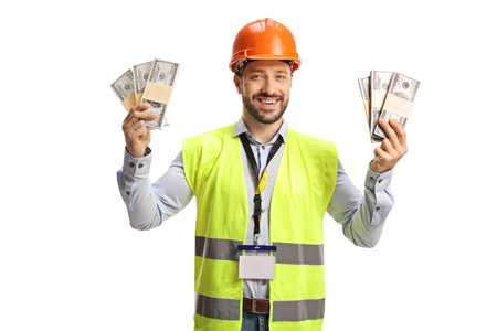 Male engineer with a safety vest and hardhat holding stacks of money and smiling isolated on white...