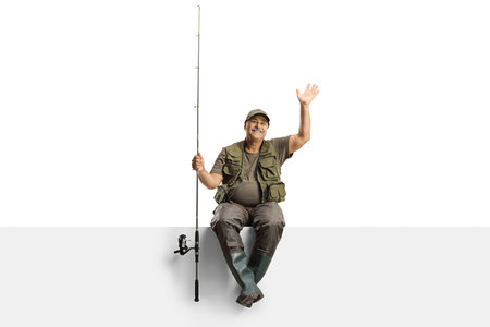 Mature fisherman with a fishing rod sitting on a blank panel and waving isolated on white background