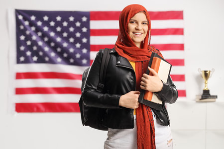 Female student with a hijab holding books and looking at camera in front of a USA flagの写真素材