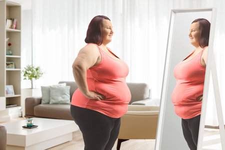 Overweight woman in sports clothes standing in front of mirror at home in a living roomの写真素材