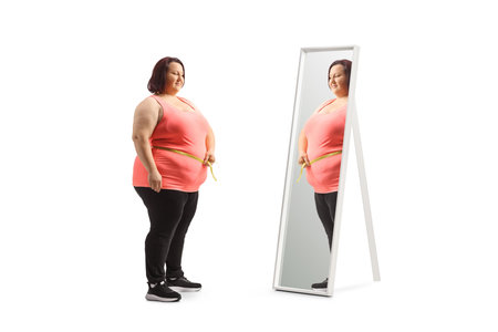 Young overweight woman measuring her waist in front of a mirror isolated on white backgroundの写真素材