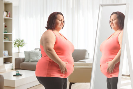 Overweight woman holding her belly and looking at a mirror at homeの写真素材