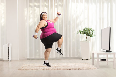 Overweight woman exercising with weights and smiling in front of tv at homeの写真素材