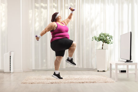 Cheerful overweight woman exercising with small dumbbells in front of tv at homeの写真素材