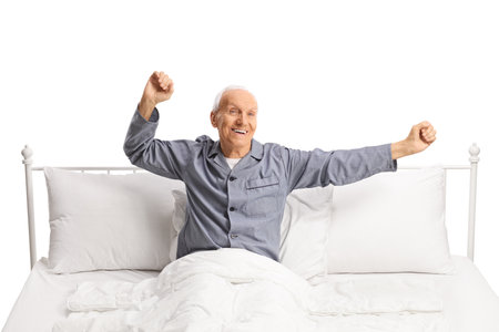 Happy elderly man stretching in a comfortable bed isolated on white backgroundの写真素材
