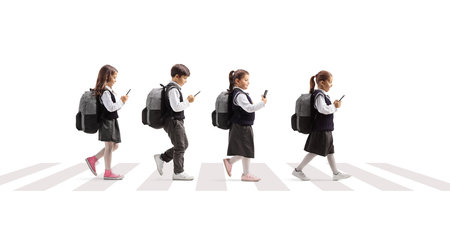 Full length profile shot of schoolchildren walking at a pedestrian crossing and using smartphones...