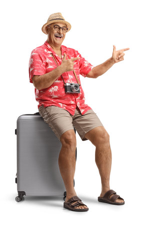 Excited male traveler sitting on a suitcase and pointing isolated on white abackgroundの写真素材