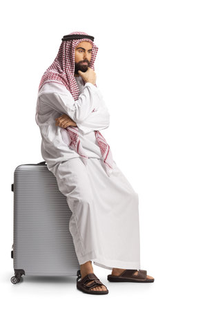 Pensive saudi arab man sitting on a suitcase isolated on white backgroundの写真素材