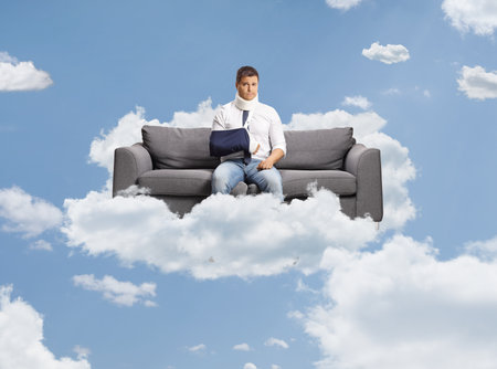 Sad man with a broken arm and neck brace sitting on a sofa floating up in the skyの写真素材
