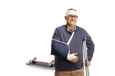 Mature man with a broken arm and bandage on head leaning on a crutch, skateboard fall injury isolated on white backgroundの写真素材
