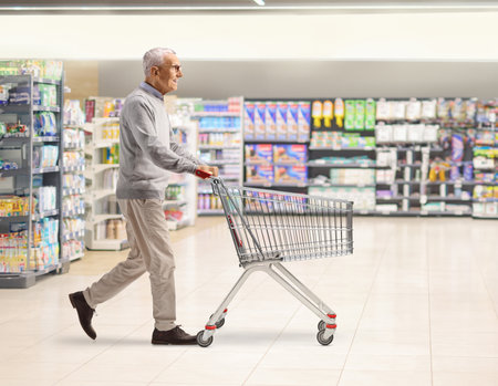 Full length profile shot of an elderly man pushing an empty shopping cart inside a supermarketの写真素材