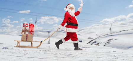 Santa Claus pulling a wooden sled with presents on a mountain and wavingの写真素材