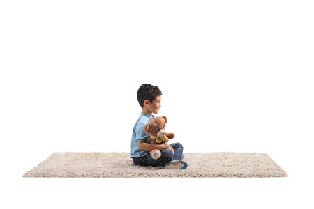 Boy holding a teddy bear and sitting on a carpet isolated on white backgroundの写真素材