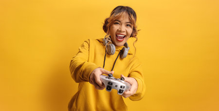 Excited asian woman with headphones holding a joystick for video games against a yellow backgroundの写真素材