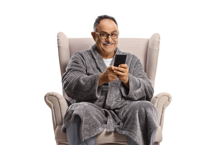 Man in a bathrobe sitting in an armchair and using a smartphone isolated on white backgroundの写真素材