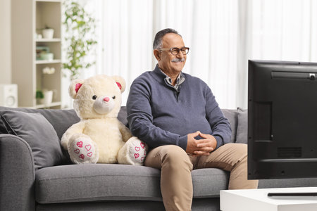 Mature man with a teddy bear sitting on a sofa and watching tv at homeの写真素材