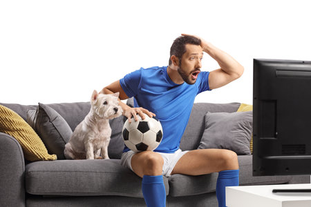 Male athlete with a dog sitting on a sofa, holding a football and watching a match on tv isolated on white backgroundの写真素材