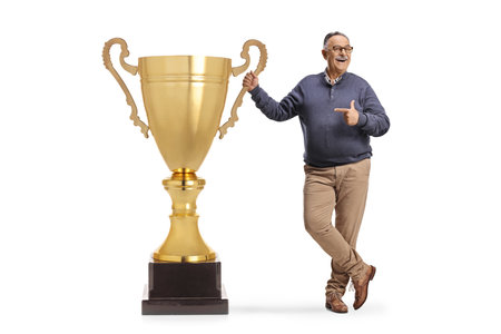 Cheerful mature man pointing at a big gold trophy cup isolated on white backgroundの写真素材